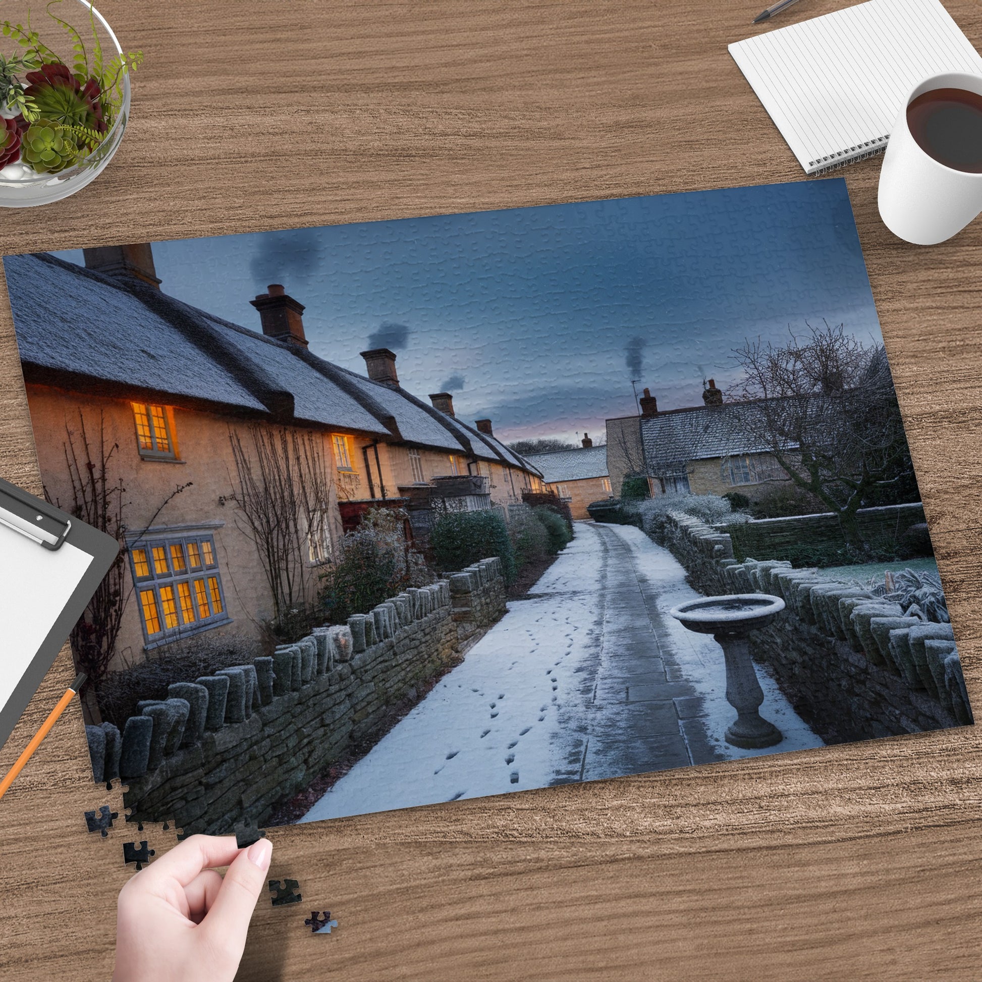 Snowy village lane with stone walls, thatched cottages, and chimney smoke at dusk.