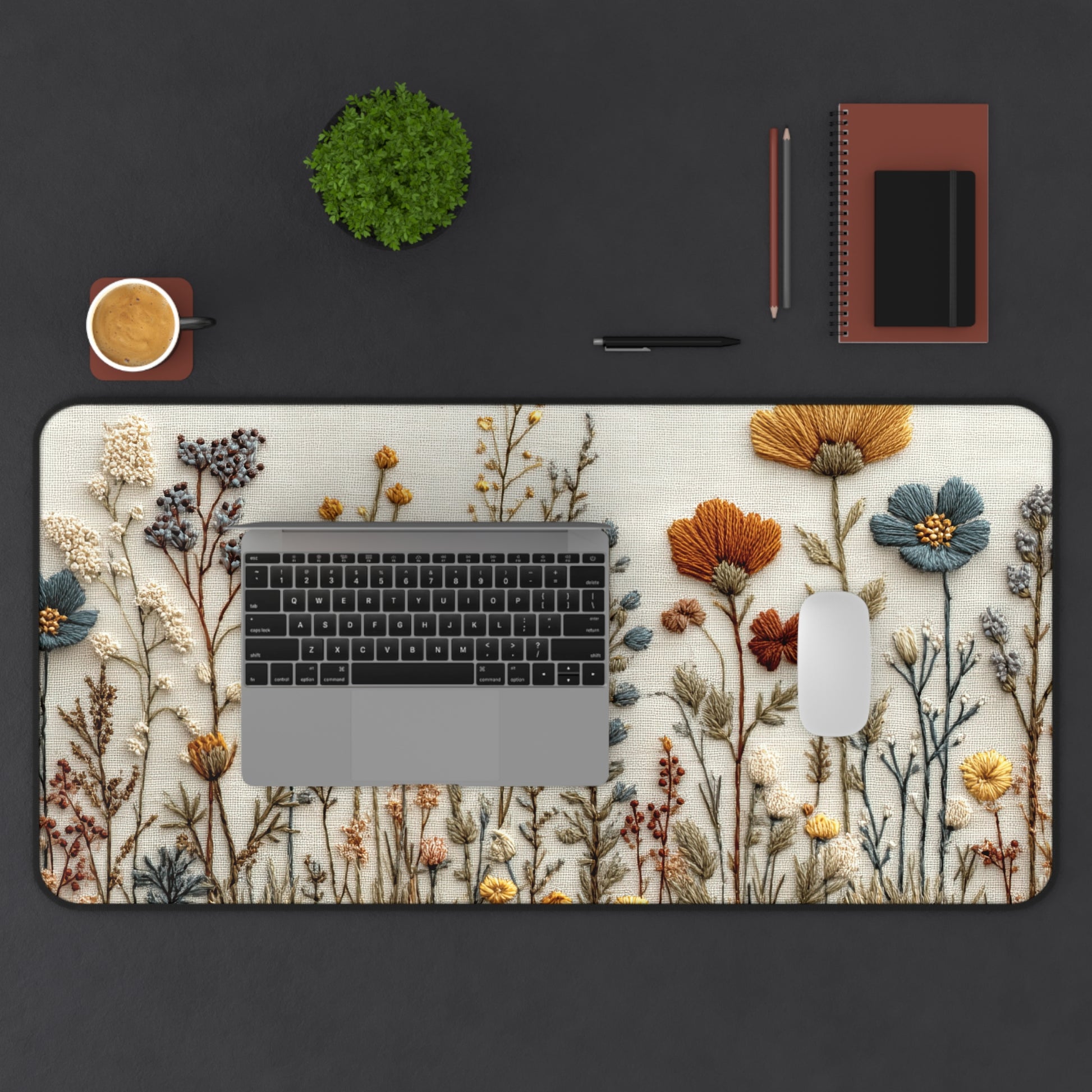 Desk setup with a floral desk mat, laptop, mouse, cup, and stationery items on a dark surface.