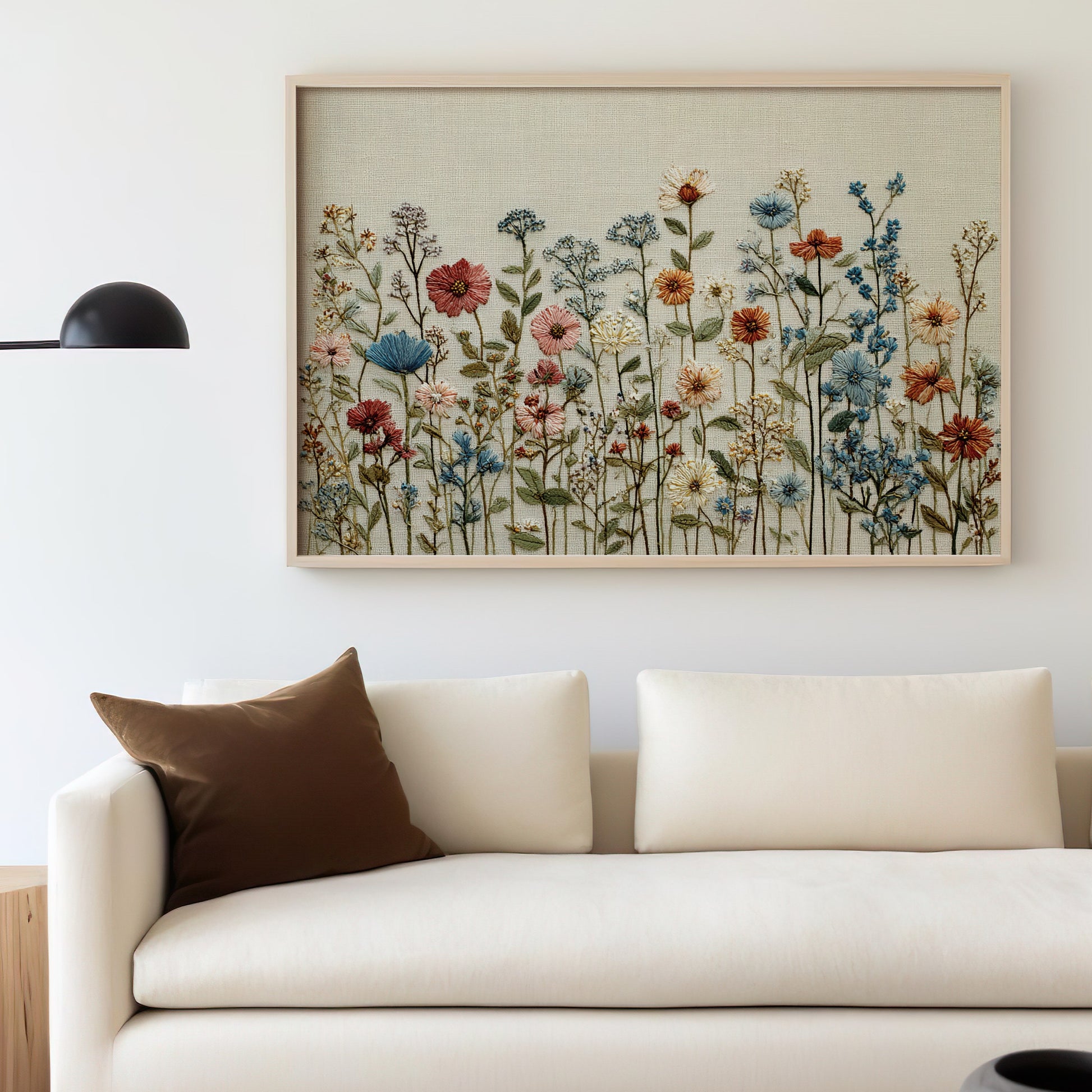 Embroidered artwork of wildflowers in a light wood frame above a cream sofa.